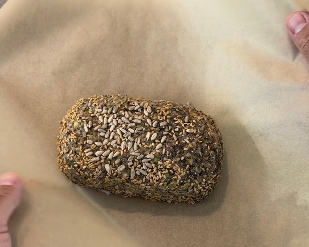 Hände halten das angehobene Backpapier mit dem geformten BROT OHNE Saatenbrotlaib